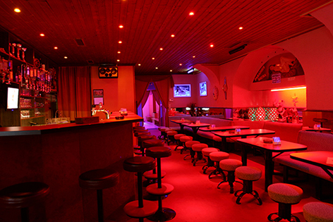 Bar mit roter Beleuchtung, Hockern und Tischen.